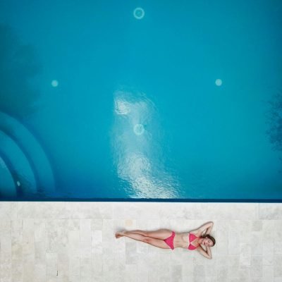 Bird's-eye view of a woman in a pink bikini relaxing by a bright blue swimming pool.
